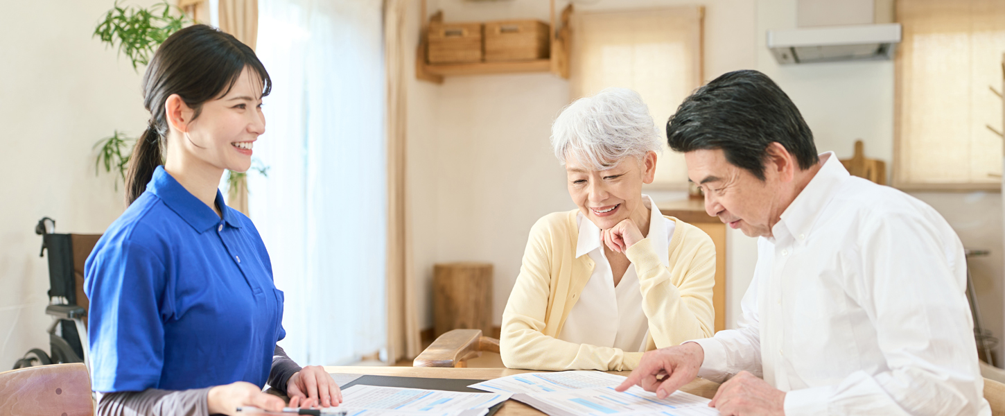 ケアプランセンターひまわりの様子：笑顔の介護士と高齢のご夫婦がテーブルで資料を見て話している